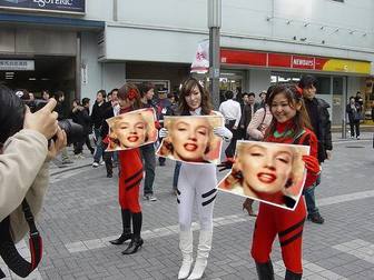 Fotomontagem em que três garotas asiáticas segurando cartazes com sua foto, na rua, com grande expectativa.